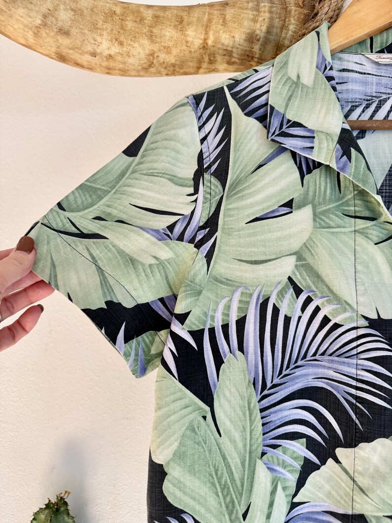 Peut inclure: Une chemise &agrave; manches courtes avec un imprim&eacute; de feuilles tropicales. La chemise pr&eacute;sente de grandes feuilles vertes avec des accents bleus et blancs sur un fond noir. Le col et la manche sont visibles.