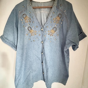 May include: Light blue denim button-down shirt with short sleeves. The shirt features gold and black embroidered embellishments and small star-shaped accents. The shirt is hanging on a wooden hanger.