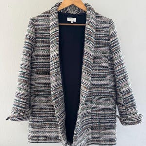 May include: A multi-colored tweed blazer with a black interior lining hangs on a wooden hanger. The blazer features a mix of colors including white, green, brown, and black. The sleeves are cuffed, and the blazer has two front pockets.