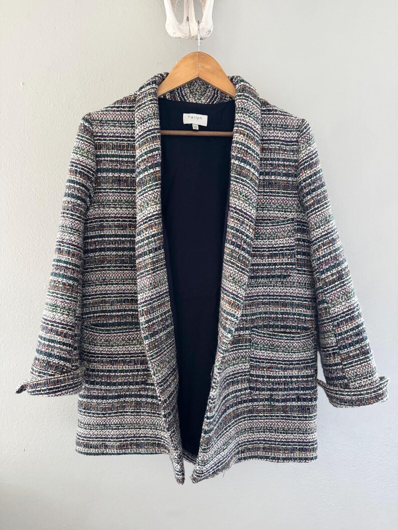 May include: A multi-colored tweed blazer with a black interior lining hangs on a wooden hanger. The blazer features a mix of colors including white, green, brown, and black. The sleeves are cuffed, and the blazer has two front pockets.