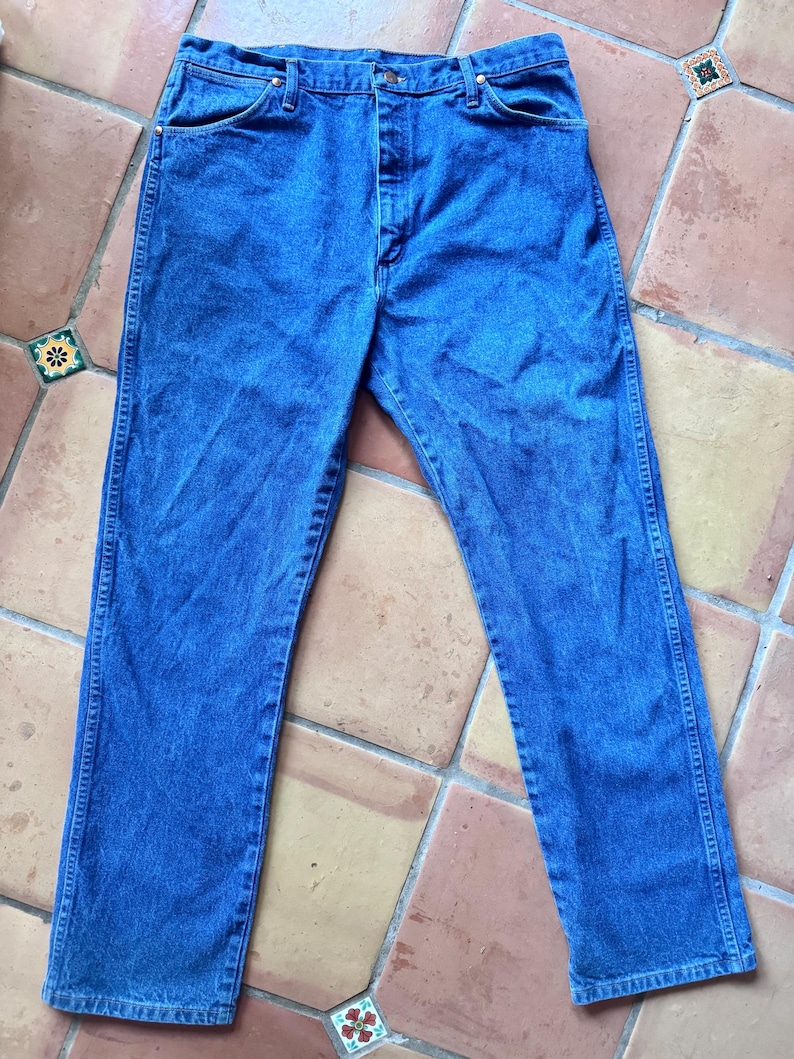 May include: A pair of medium-wash blue denim jeans featuring a classic five-pocket design, a button fly closure, and copper-toned rivets at the pocket corners. The jeans have a straight-leg cut with visible stitching along the seams and waistband.