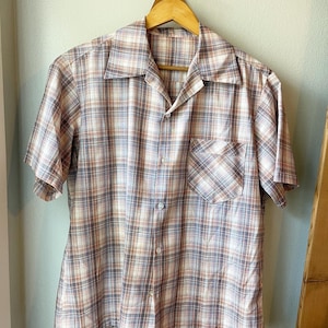 May include: A short-sleeved button-down shirt with a plaid pattern in shades of brown, blue, and white. The shirt has a collared neckline and a single chest pocket.