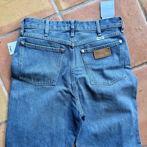 May include: Blue denim jeans with the Wrangler brand label on the back pocket. The jeans have a classic five-pocket design, copper rivets, and a straight leg fit. A price tag is attached to the jeans.
