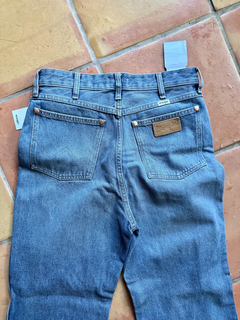 May include: Blue denim jeans with the Wrangler brand label on the back pocket. The jeans have a classic five-pocket design, copper rivets, and a straight leg fit. A price tag is attached to the jeans.