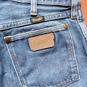 K&ouml;nnte beinhalten: Nahaufnahme einer blauen Jeans mit einem Wrangler-Etikett auf der Ges&auml;&szlig;tasche. Die Jeans hat ein klassisches F&uuml;nf-Taschen-Design mit kupferfarbenen Nieten und N&auml;hten. Das Wrangler-Logo ist auf der Ges&auml;&szlig;tasche und am Bund zu sehen.