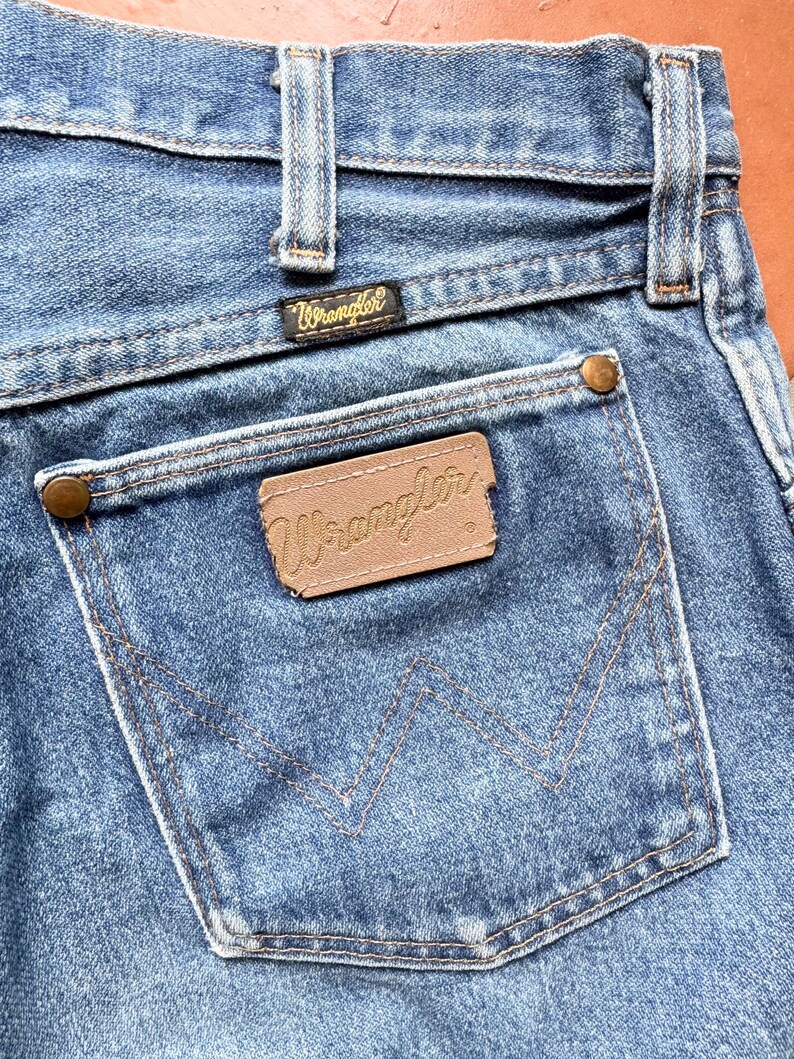 K&ouml;nnte beinhalten: Nahaufnahme einer blauen Jeans mit einem Wrangler-Etikett auf der Ges&auml;&szlig;tasche. Die Jeans hat ein klassisches F&uuml;nf-Taschen-Design mit kupferfarbenen Nieten und N&auml;hten. Das Wrangler-Logo ist auf der Ges&auml;&szlig;tasche und am Bund zu sehen.