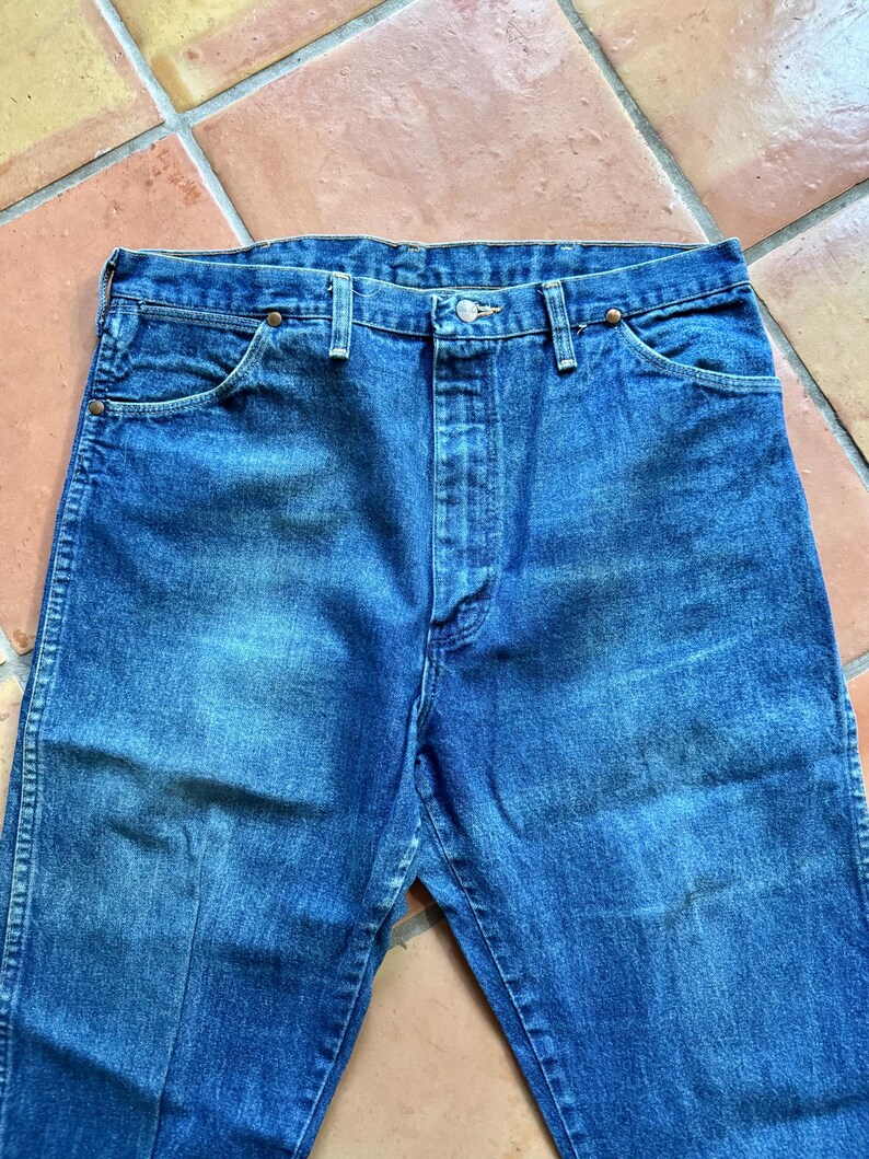 May include: Close-up of blue denim jeans. The jeans have a button closure and pockets. The denim has a faded appearance, with lighter areas on the thighs and knees. The jeans are a classic style.