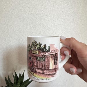 May include: White ceramic mug featuring a detailed illustration of the OK Corral in Tombstone, Arizona. The artwork includes a brick building, a stagecoach, and the text "OK CORRAL" and "Tombstone, Az.". The mug has a white handle.