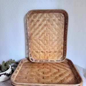 May include: A stack of rectangular woven bamboo trays. The trays have a natural light brown color and a woven pattern. The trays are stacked on top of each other, with one tray standing upright in the background.
