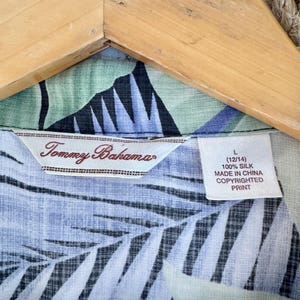 Peut inclure: Gros plan sur une chemise en soie avec un motif de feuilles tropicales dans les tons de bleu, vert et noir. L'&eacute;tiquette indique "Tommy Bahama" et comprend une &eacute;tiquette de taille L (12/14). La chemise est en 100% soie et fabriqu&eacute;e en Chine.