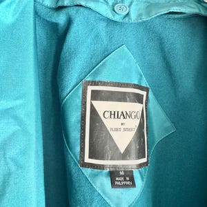 May include: Close-up of a teal jacket with a label that reads "CHIANGO BY FLEET STREET." The label is black and white with a diamond shape. The jacket has a button and a diamond-shaped patch.