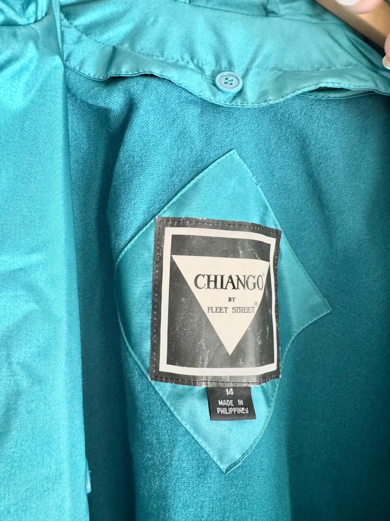 May include: Close-up of a teal jacket with a label that reads "CHIANGO BY FLEET STREET." The label is black and white with a diamond shape. The jacket has a button and a diamond-shaped patch.