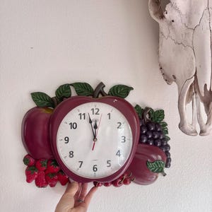 May include: A decorative wall clock shaped like an apple, with a white clock face and black numbers. The clock is surrounded by a cluster of red and purple fruits and green leaves. The clock is being held by a person.