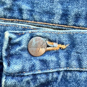 May include: Close-up of blue denim jeans with a copper-coloured button. The button is embossed with the word "Wrangler". The jeans have visible stitching in a contrasting tan colour.
