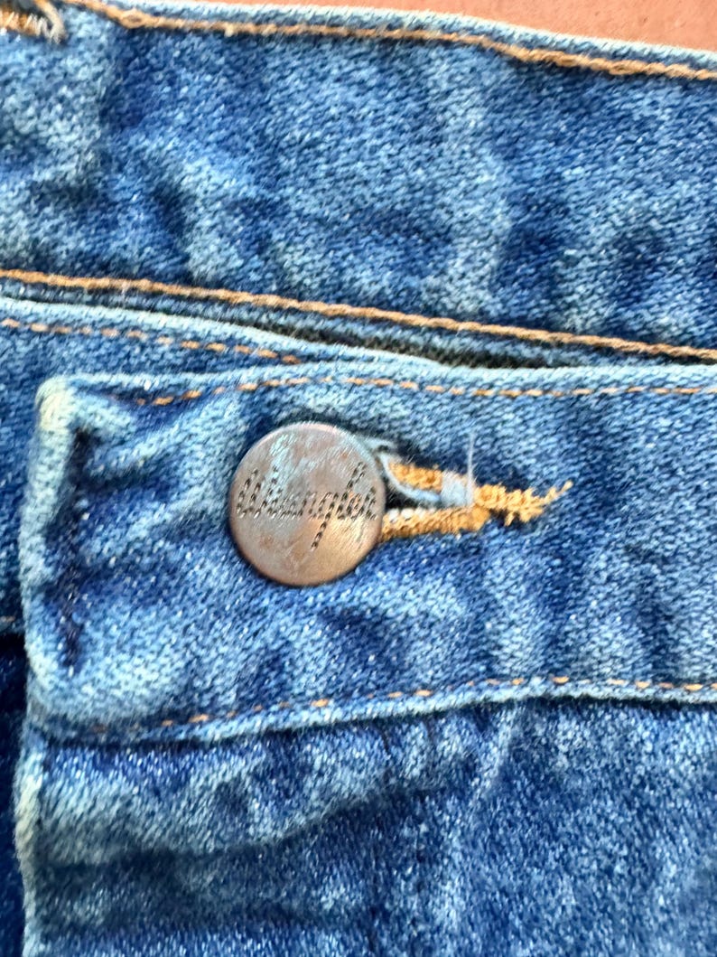 May include: Close-up of blue denim jeans with a copper-coloured button. The button is embossed with the word "Wrangler". The jeans have visible stitching in a contrasting tan colour.