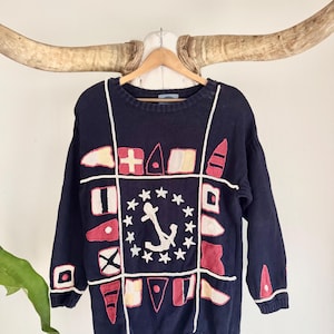 May include: Navy blue sweater with nautical flag and anchor design. The sweater features a crew neck, long sleeves, and white trim. The anchor is surrounded by stars. The flags are red, white, and beige.