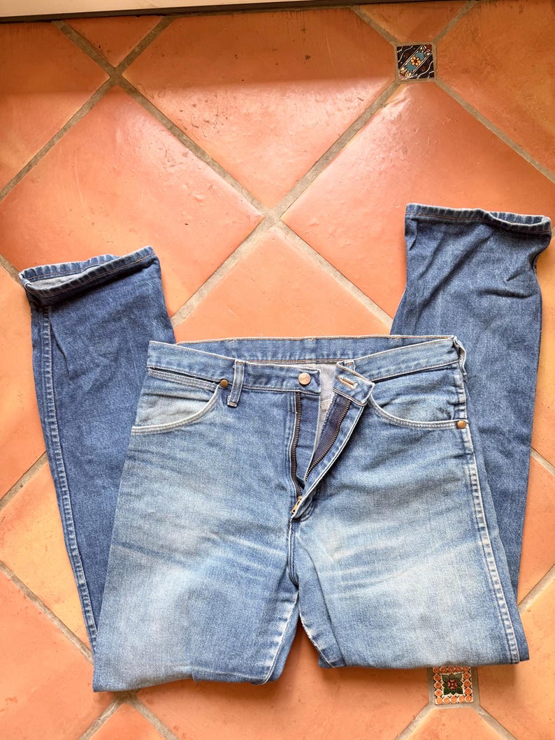 K&ouml;nnte beinhalten: Hellblaue Jeans aus Denim mit klassischem F&uuml;nf-Taschen-Design. Die Jeans liegen flach, wobei die Vorderseite mit teilweise ge&ouml;ffnetem Rei&szlig;verschluss zu sehen ist. Der Stoff hat ein verwaschenes Aussehen.