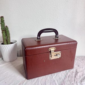 May include: A rectangular, dark brown, textured vanity case featuring a dark brown top handle and a central metallic latch mechanism. The case has a structured, boxy shape with visible hardware details on the front closure and handle mounts.