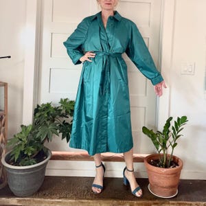 May include: Teal-colored trench coat with a button-up front, long sleeves, and a matching belt. The coat has a classic collar and pockets. The model is wearing blue heels. The coat is knee-length and made of a shiny material.