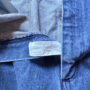 May include: Blue denim jeans featuring a white interior tag with the Wrangler logo, care instructions, and the text Made in USA 100% Cotton. The jeans have a metal rivet at the pocket corner and visible stitching along the waistband and fly.