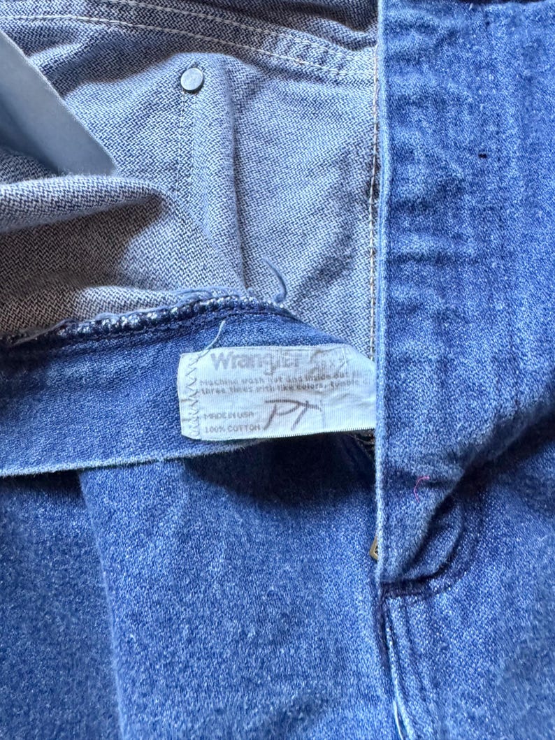 May include: Blue denim jeans featuring a white interior tag with the Wrangler logo, care instructions, and the text Made in USA 100% Cotton. The jeans have a metal rivet at the pocket corner and visible stitching along the waistband and fly.