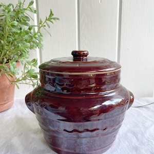 May include: A brown ceramic crock with a lid. The crock has a wavy design around the middle and a floral design on the sides. The lid has a knob on top.