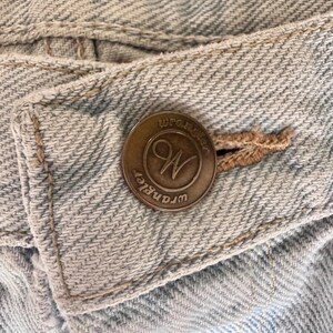 May include: Close-up of a light blue denim fabric with a Wrangler button. The button is bronze-colored with the Wrangler logo in a circular design. The denim has visible stitching and a classic, worn look.