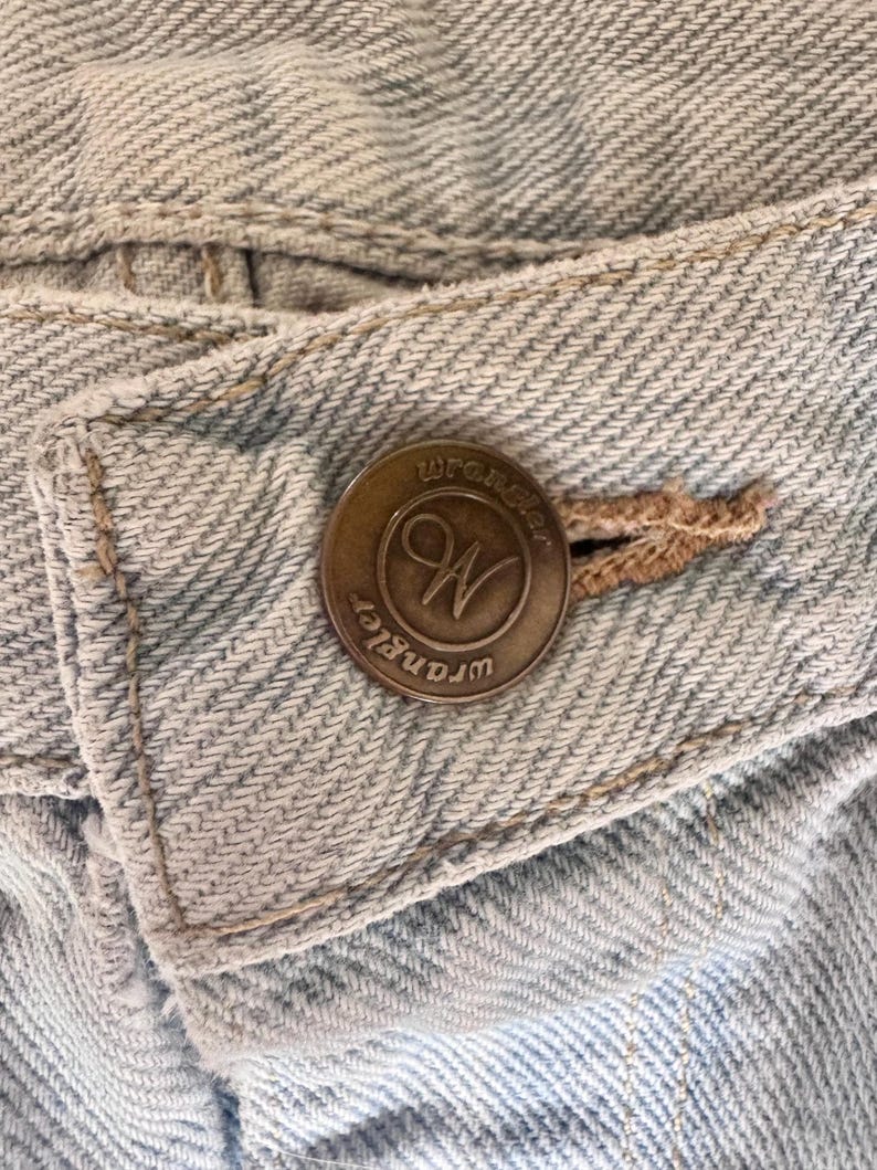 May include: Close-up of a light blue denim fabric with a Wrangler button. The button is bronze-colored with the Wrangler logo in a circular design. The denim has visible stitching and a classic, worn look.