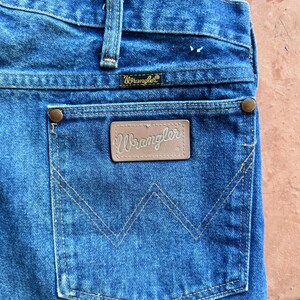 May include: Close-up of blue denim jeans showing the back pocket with the Wrangler logo. The pocket has a distinctive stitched 'W' design. The jeans have a classic, worn appearance, with visible stitching and rivets.