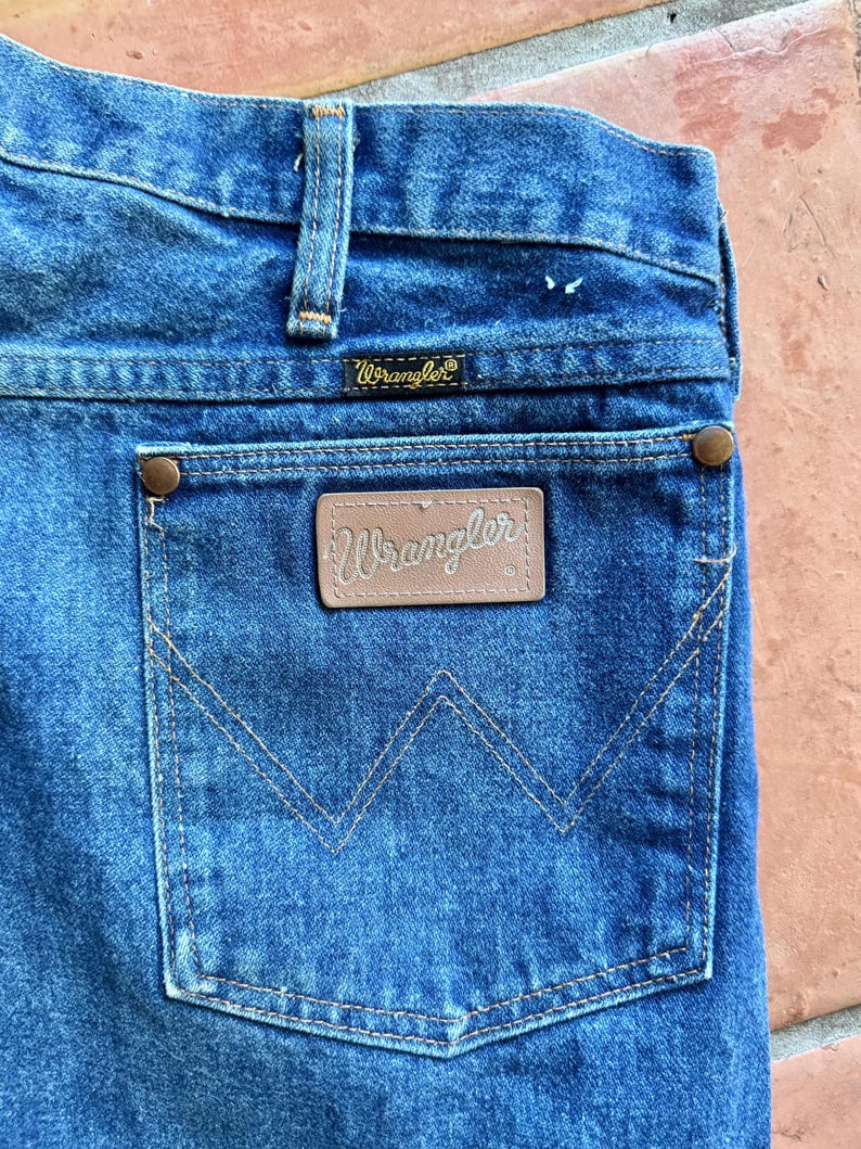 May include: Close-up of blue denim jeans showing the back pocket with the Wrangler logo. The pocket has a distinctive stitched 'W' design. The jeans have a classic, worn appearance, with visible stitching and rivets.