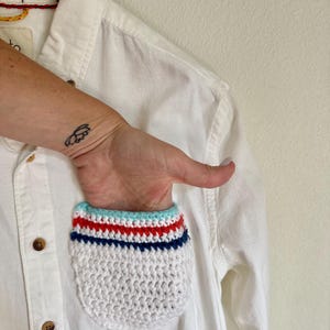 May include: White button-down shirt with a crocheted pocket. The pocket features a white base with horizontal stripes in red, blue, and light blue. The shirt is hanging on a wooden hanger.