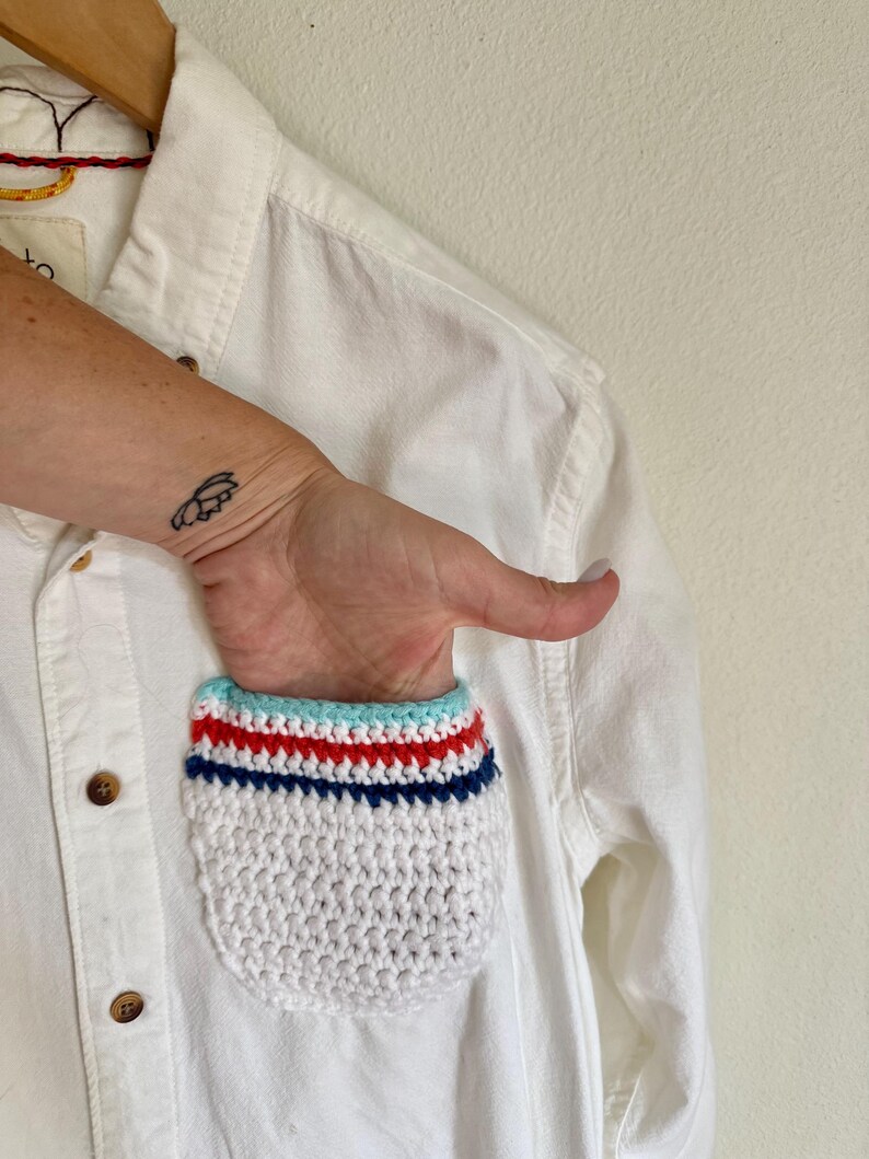 May include: White button-down shirt with a crocheted pocket. The pocket features a white base with horizontal stripes in red, blue, and light blue. The shirt is hanging on a wooden hanger.