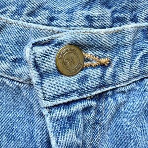 May include: Close-up of light blue denim jeans with a brass button. The button is embossed with the word "Wrangler" and the letters "W" and "N" inside a circle, with the word "America" around the edge. The jeans have a classic five-pocket design.