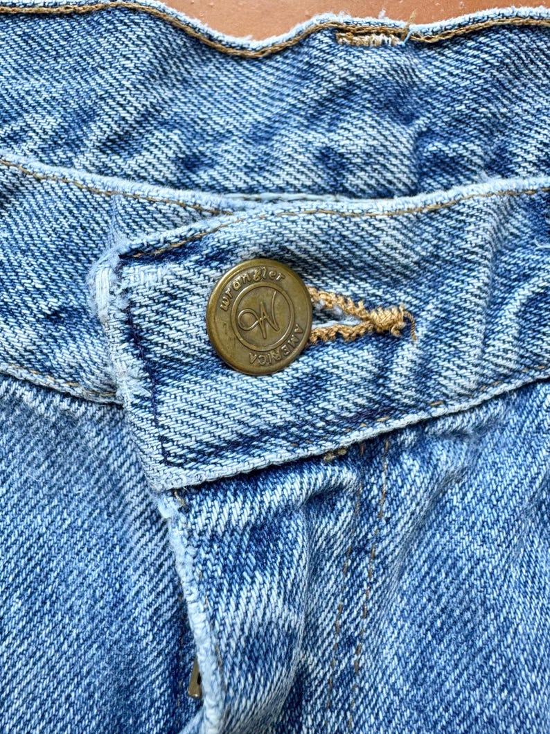 May include: Close-up of light blue denim jeans with a brass button. The button is embossed with the word "Wrangler" and the letters "W" and "N" inside a circle, with the word "America" around the edge. The jeans have a classic five-pocket design.