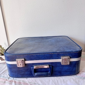 May include: A vintage blue suitcase with silver-colored hardware, including two latches and a handle. The suitcase has a white stripe around the middle and is sitting on a patterned fabric.