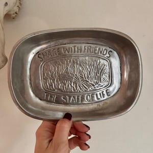 May include: A silver-toned, oval-shaped tray with the embossed text "SHARE WITH FRIENDS" and "THE STAFF OF LIFE." The tray features a central embossed design of tall grass or reeds. The tray is held by a hand with dark nail polish.