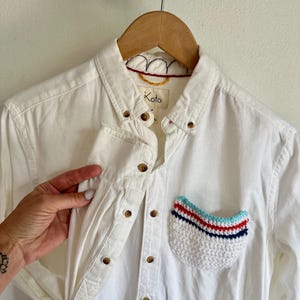 May include: White button-down shirt with a crocheted pocket. The pocket features a white base with red, blue, and light blue stripes. The shirt has wooden buttons and the brand name "Koto" is visible.