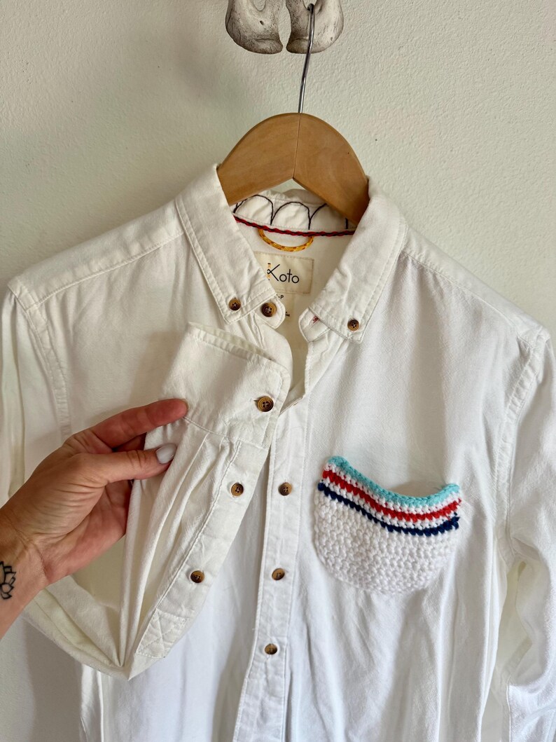 May include: White button-down shirt with a crocheted pocket. The pocket features a white base with red, blue, and light blue stripes. The shirt has wooden buttons and the brand name "Koto" is visible.