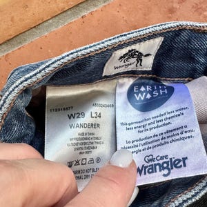 May include: Close-up of the inside label of Wrangler jeans. The label shows the size W29 L34, the word "Wanderer", and the "Earth Wash" logo. The label also includes washing instructions and the Wrangler logo.