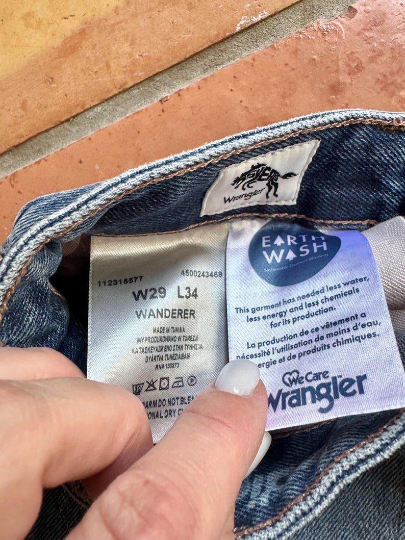 May include: Close-up of the inside label of Wrangler jeans. The label shows the size W29 L34, the word "Wanderer", and the "Earth Wash" logo. The label also includes washing instructions and the Wrangler logo.