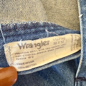 May include: Close-up of a blue denim Wrangler jeans tag. The tag reads "Wrangler" with the size 35 x 32. The tag also includes washing instructions and "MADE IN USA" with 100% cotton.