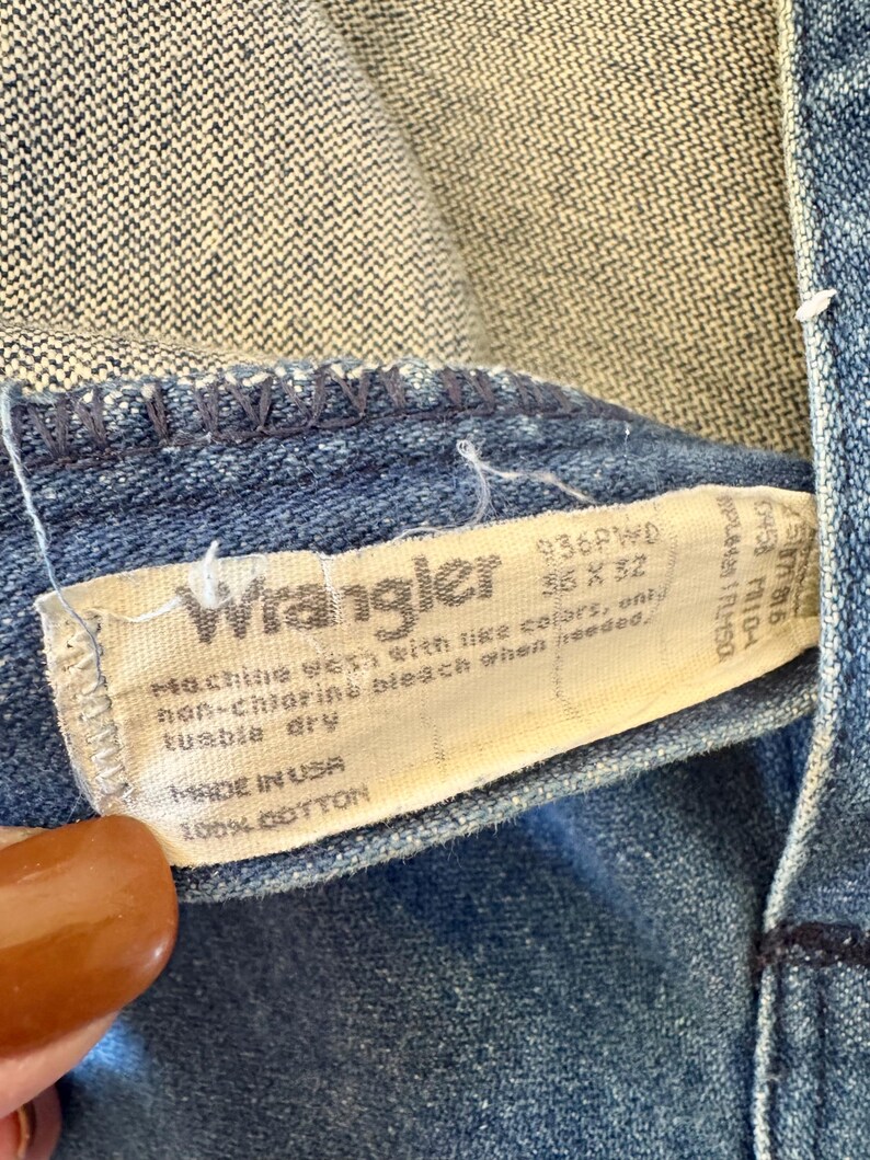 May include: Close-up of a blue denim Wrangler jeans tag. The tag reads "Wrangler" with the size 35 x 32. The tag also includes washing instructions and "MADE IN USA" with 100% cotton.