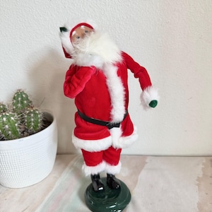 May include: A festive Santa Claus figurine in a red velvet suit with white faux fur trim. The figure has a white beard, black boots, and a green base. A small cactus in a white pot is in the background. Christmas decor.