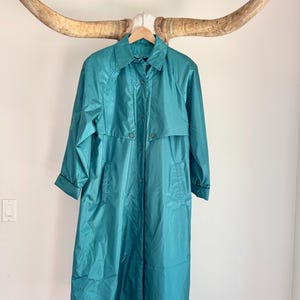 May include: Teal-colored vintage raincoat with a collared neckline and button-down closure. The long coat features a classic design with a mid-length hem and long sleeves. The coat is hanging on a wooden hanger.