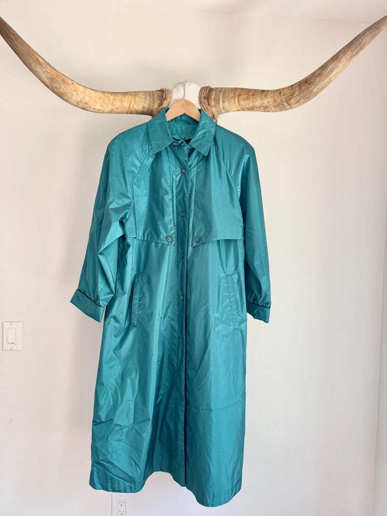 May include: Teal-colored vintage raincoat with a collared neckline and button-down closure. The long coat features a classic design with a mid-length hem and long sleeves. The coat is hanging on a wooden hanger.