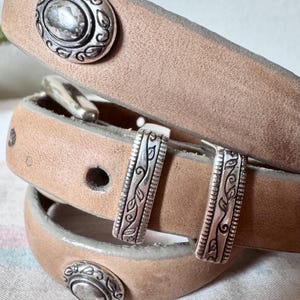 May include: A light brown leather belt with silver metal detailing. The belt has decorative oval studs and rectangular buckles with leaf patterns. The belt is rolled up, displaying the buckle and stud details.