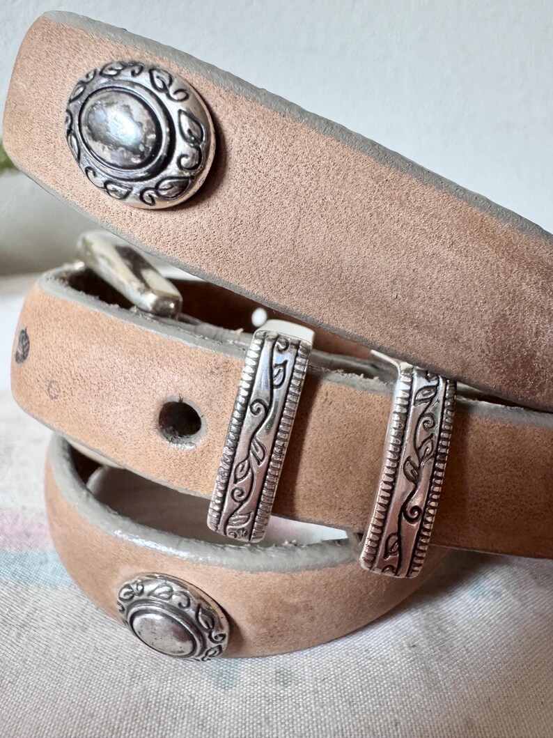 May include: A light brown leather belt with silver metal detailing. The belt has decorative oval studs and rectangular buckles with leaf patterns. The belt is rolled up, displaying the buckle and stud details.