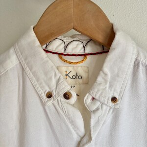May include: Close-up of a white button-down shirt hanging on a wooden hanger. The shirt features brown buttons, a collar, and a label that reads "Koto". The collar has decorative stitching in red, black, and yellow.