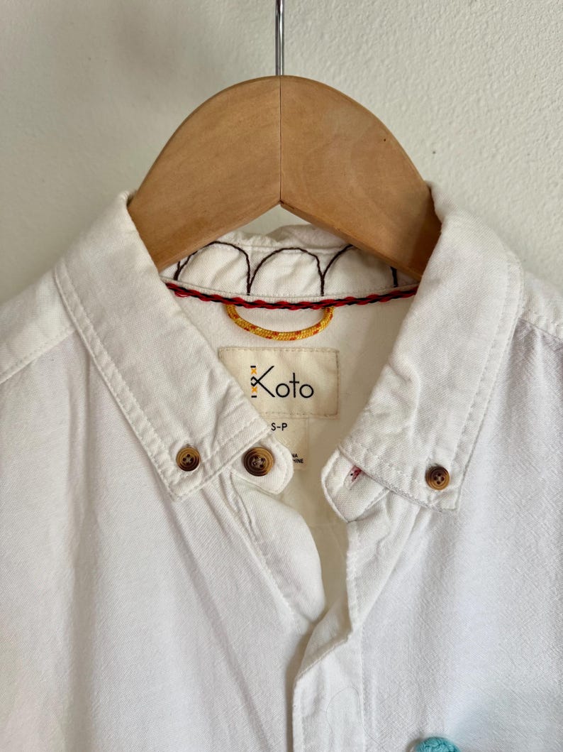 May include: Close-up of a white button-down shirt hanging on a wooden hanger. The shirt features brown buttons, a collar, and a label that reads "Koto". The collar has decorative stitching in red, black, and yellow.