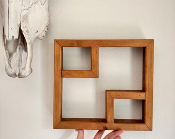 May include: A square, wooden wall shelf with a geometric design. The shelf is made of light brown wood and has an open, box-like structure with multiple compartments. A cow skull is visible in the background.